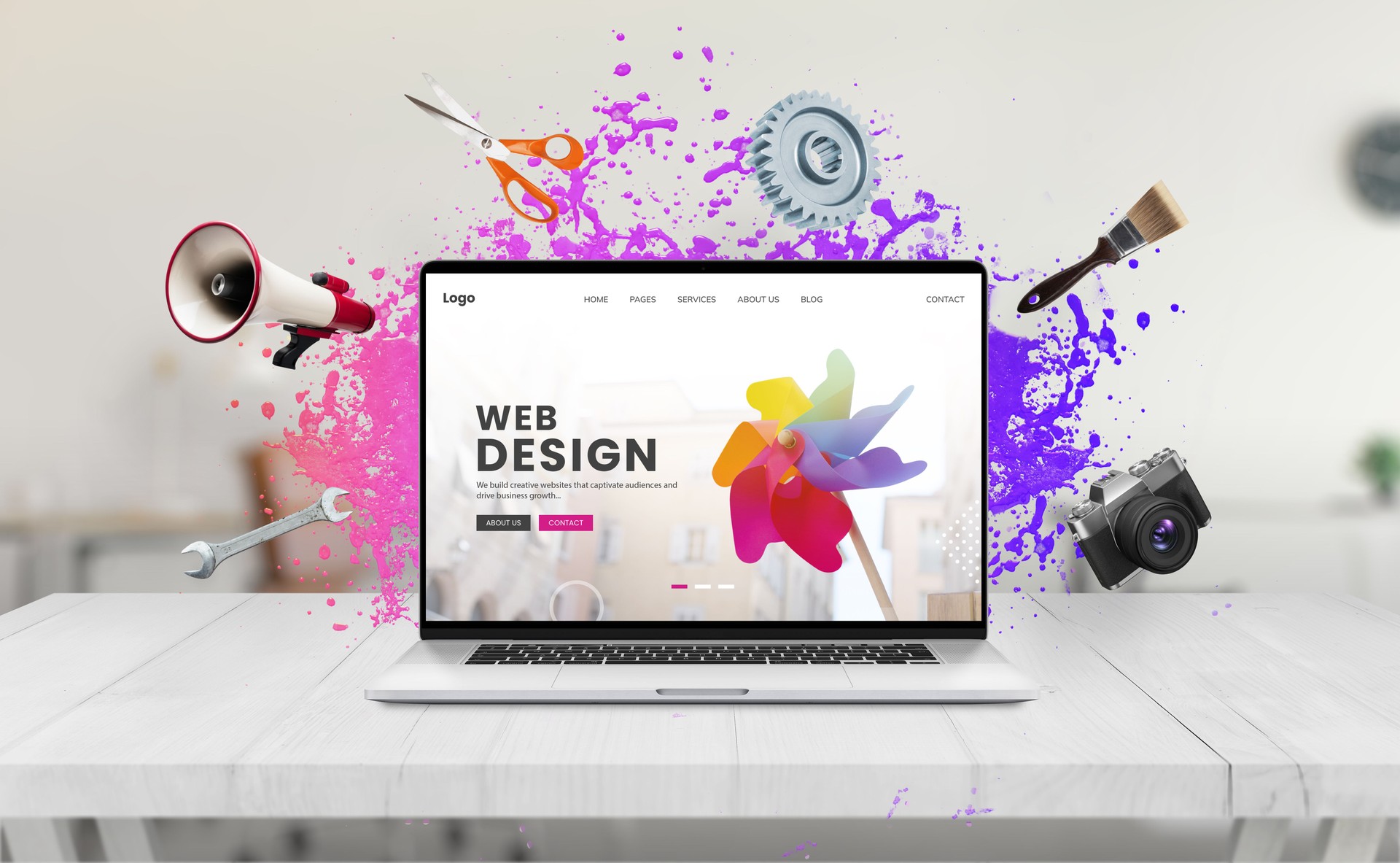 Laptop on a desk displaying a web design concept page featuring a colorful water splash, surrounded by creative tools like a brush, megaphone, wrench, camera, scissors, and gear